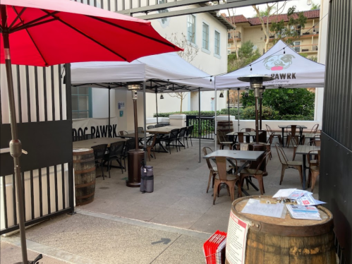 Outdoor dining area with red umbrellas, tables, and tents labeled "DOG PAWRK," surrounded by greenery.
