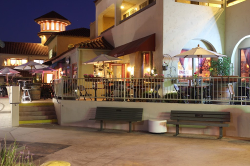 A cozy outdoor dining area with tables, umbrellas, and benches, illuminated at night near a building with a tower.