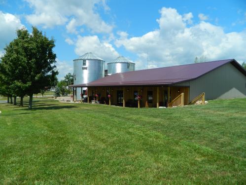 A modern building with a metal roof, surrounded by green grass and trees, next to two silver silos under a blue sky.