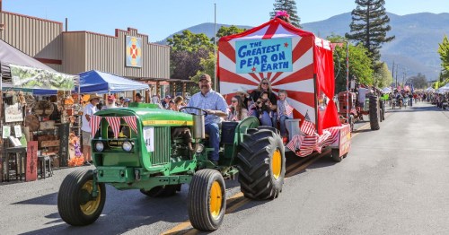 A green tractor pulls a festive float reading "The Greatest Pear on Earth" during a lively parade in a small town.