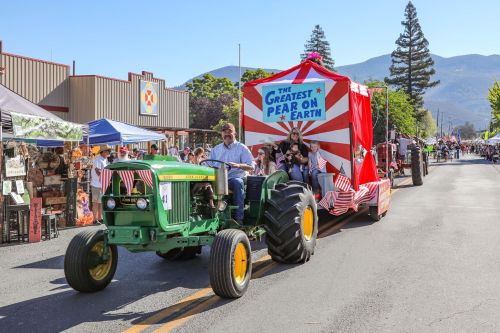 A green tractor leads a festive float decorated with red and white, promoting "The Greatest Pear on Earth" at a parade.