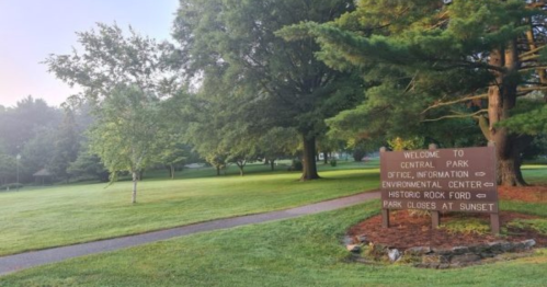 A sign welcomes visitors to Central Park, surrounded by trees and a grassy area, with a path leading through the park.