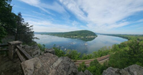 Panoramic view of a river surrounded by lush greenery and hills under a blue sky with scattered clouds.