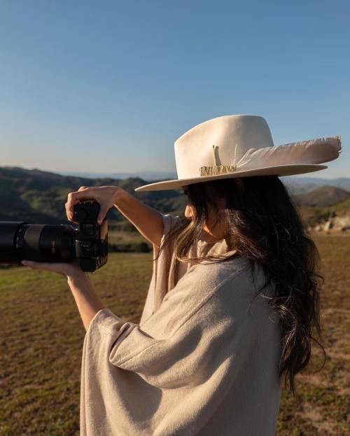 A woman in a wide-brimmed hat holds a camera, capturing a scenic landscape under a clear blue sky.
