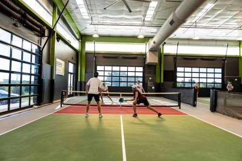 Two players engage in a game of pickleball indoors, with a net and large windows in the background.