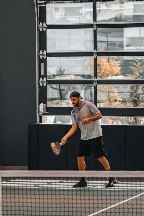 A man prepares to serve on a pickleball court, wearing a gray shirt and black shorts, with a garage door backdrop.