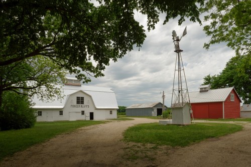 A scenic farm with a white barn, red barn, windmill, and cloudy sky, surrounded by green trees and a dirt path.