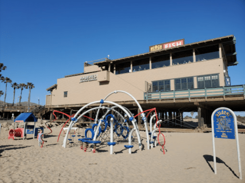 A beach playground with colorful equipment in front of a restaurant on a pier, with palm trees and clear blue skies.