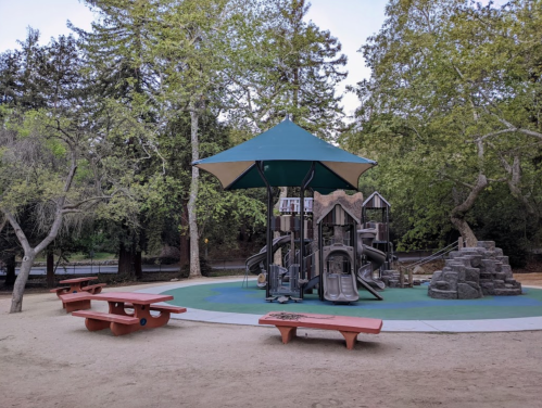Playground with slides and climbing structures under a large green canopy, surrounded by trees and picnic tables.