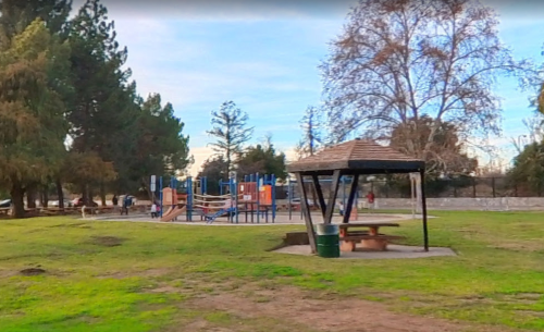 A playground with climbing structures and a shaded picnic area in a grassy park surrounded by trees.