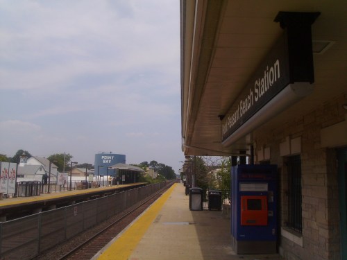 A train station platform with a sign reading "Tennessee Beach Station," featuring a ticket machine and empty tracks.