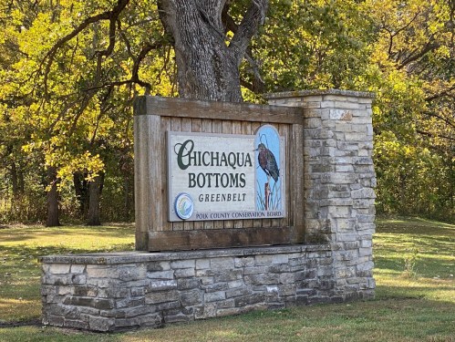 Sign for Chichaqua Bottoms Greenbelt, featuring a bird illustration and surrounded by trees in a natural setting.