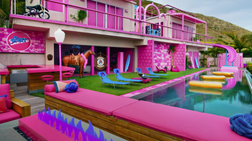 A vibrant pink-themed pool area with lounge chairs, a water slide, and a horse statue near a modern building.