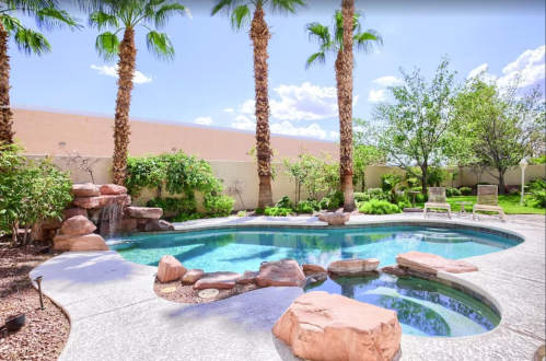 A serene backyard with a pool surrounded by palm trees and lush greenery, featuring rock formations and lounge chairs.