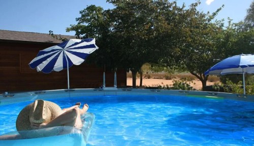 A person relaxes on a float in a blue pool, surrounded by trees and striped umbrellas on a sunny day.