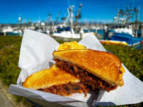 A close-up of a grilled sandwich filled with pulled pork, served with macaroni and cheese, with a harbor in the background.