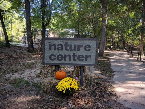 Sign for a nature center surrounded by trees, with a pumpkin and flowers at its base.