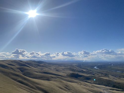 A bright sun shines over rolling hills and a winding river, with fluffy clouds scattered in a blue sky.