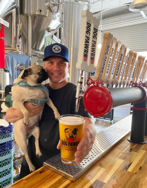 A man holds a pint of beer in a brewery while cuddling a pug dog. Beer taps labeled "DOG PAW" are in the background.