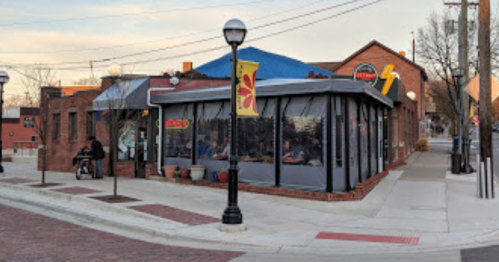 A brick restaurant with large windows, outdoor seating, and a colorful banner, located on a street corner.