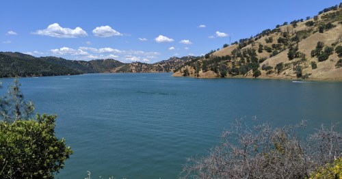 A serene lake surrounded by rolling hills and trees under a clear blue sky with scattered clouds.