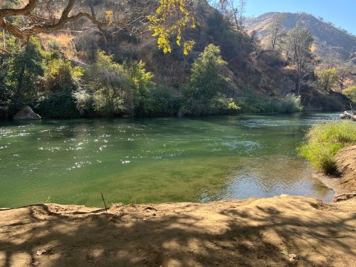 A serene river flows through a lush landscape, surrounded by hills and trees under a clear blue sky.