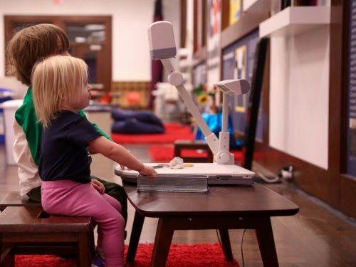 Two young children sit at a small table, focused on a projector in a cozy, colorful learning environment.