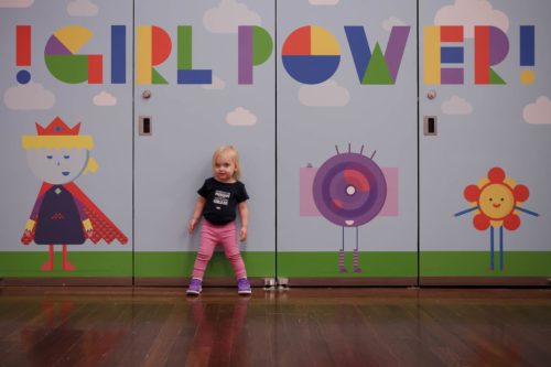 A young girl stands in front of colorful "Girl Power!" murals featuring playful characters on a wall.