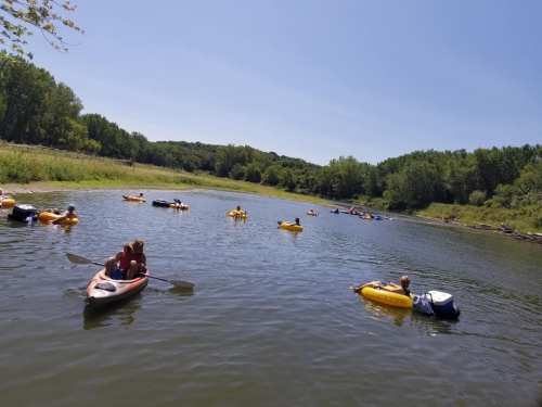 A sunny day on a river with people kayaking and floating in yellow inflatable boats surrounded by greenery.