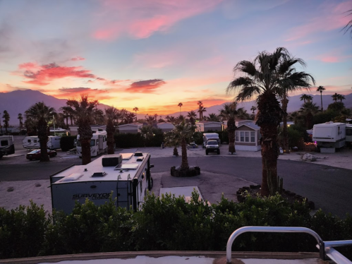 Sunset over a desert RV park, with palm trees and parked RVs silhouetted against a colorful sky.