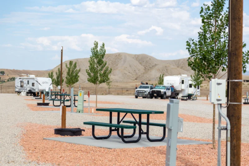 A gravel campsite with picnic tables, trees, and RVs in the background against a hilly landscape under a blue sky.