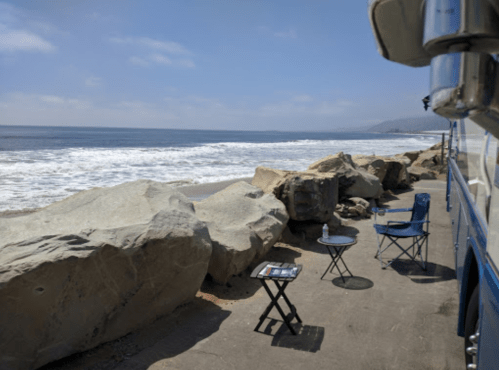 A peaceful beach scene with large rocks, a small table, a chair, and waves gently lapping at the shore.