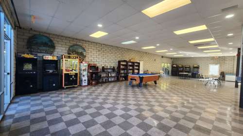 A spacious room with arcade machines, a pool table, bookshelves, and tables, featuring a checkered floor and bright lighting.