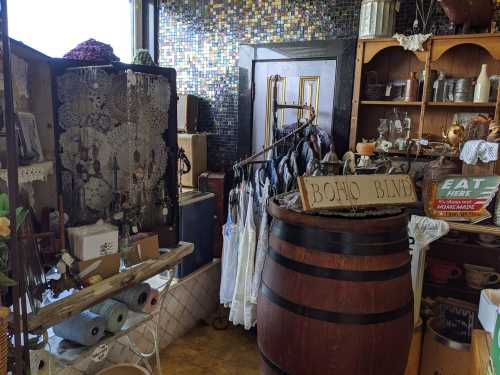 A cozy vintage shop interior featuring lace items, clothing racks, and a wooden barrel with a sign reading "BOHO BLVD."