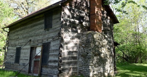 A rustic log cabin with a stone chimney, surrounded by greenery and trees in a peaceful outdoor setting.