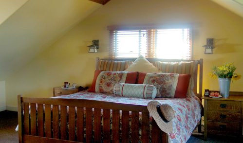Cozy attic bedroom with a wooden bed, floral bedding, and natural light from windows. Decorated with a vase of flowers.