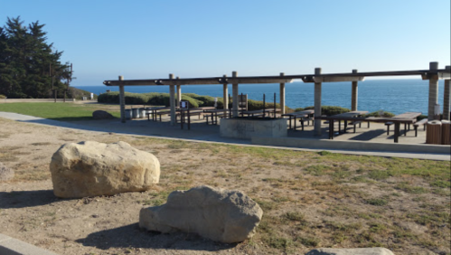 Outdoor picnic area with tables and ocean view, surrounded by grass and large rocks under a clear blue sky.