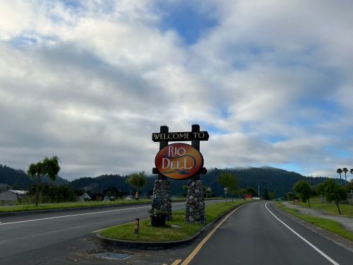 Welcome sign for Rio Dell, surrounded by trees and mountains under a cloudy sky.