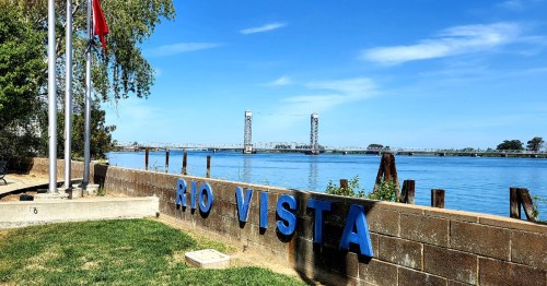 Sign reading "Rio Vista" by the water, with a bridge in the background and clear blue skies above.