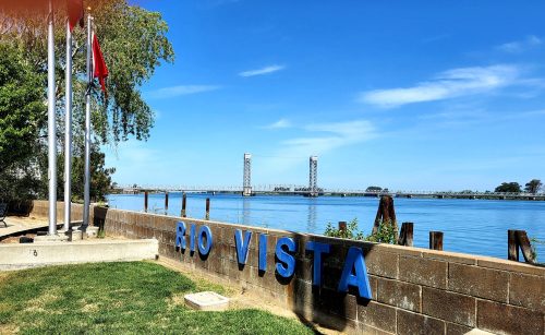 A scenic view of the Rio Vista sign by the water, with a bridge in the background and clear blue skies.