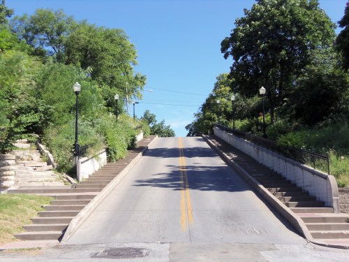 A wide, tree-lined street slopes upward, flanked by stairs and lampposts under a clear blue sky.