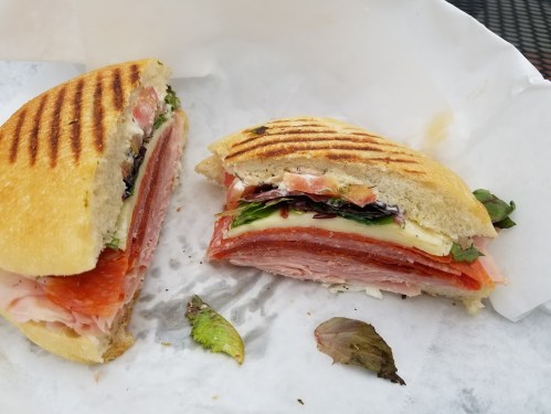 A toasted sandwich cut in half, filled with layers of meats, cheese, and greens, resting on white parchment paper.