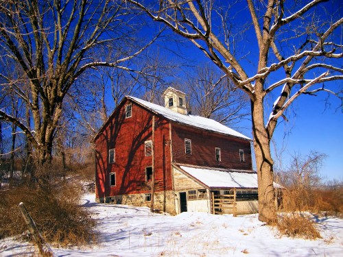 A red barn with a snow-covered roof, surrounded by bare trees and a clear blue sky.