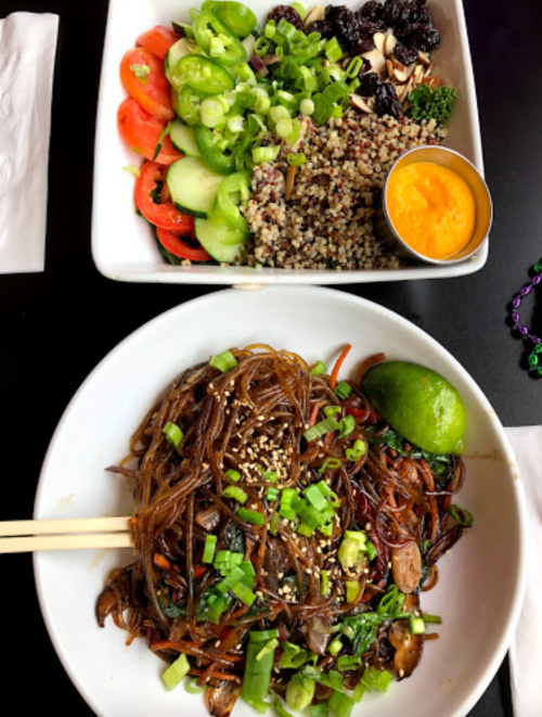 A bowl of stir-fried noodles topped with green onions and sesame seeds, alongside a fresh salad and quinoa.