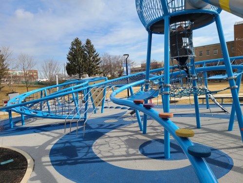 A colorful playground with blue structures, climbing nets, and slides, set against a cloudy sky.