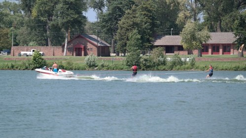 A boat pulls two water skiers across a calm lake, with trees and buildings in the background.