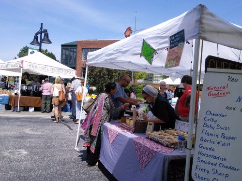 A bustling farmers market with vendors selling cheese and coffee, and shoppers browsing under sunny skies.