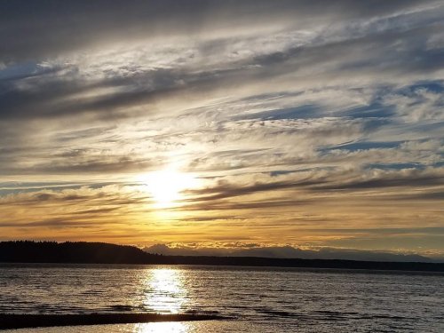 A serene sunset over a calm body of water, with vibrant clouds and distant mountains silhouetted against the sky.