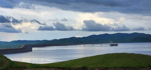 A serene landscape featuring a calm lake, distant mountains, and a bridge extending over the water under a cloudy sky.