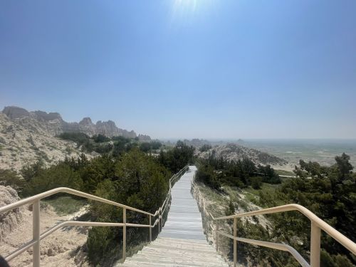 A wooden walkway leads through greenery, with rugged rock formations and a clear blue sky in the background.